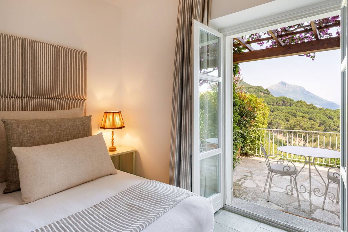 A bedroom with a bed, pillows, a nightstand, a lamp, and a view of a balcony with a table and chairs.