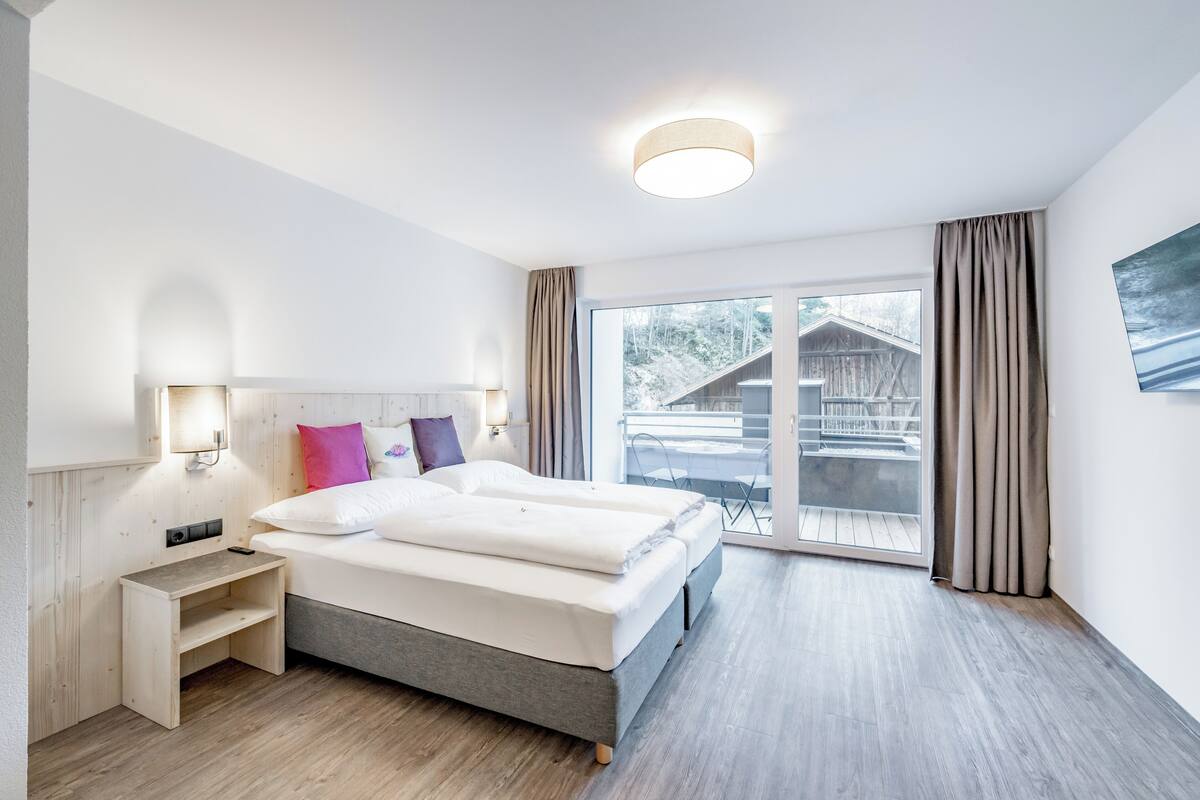 A modern hotel room with a large bed, bedside tables, a nightstand, and a view of a wooden cabin through a sliding glass door.