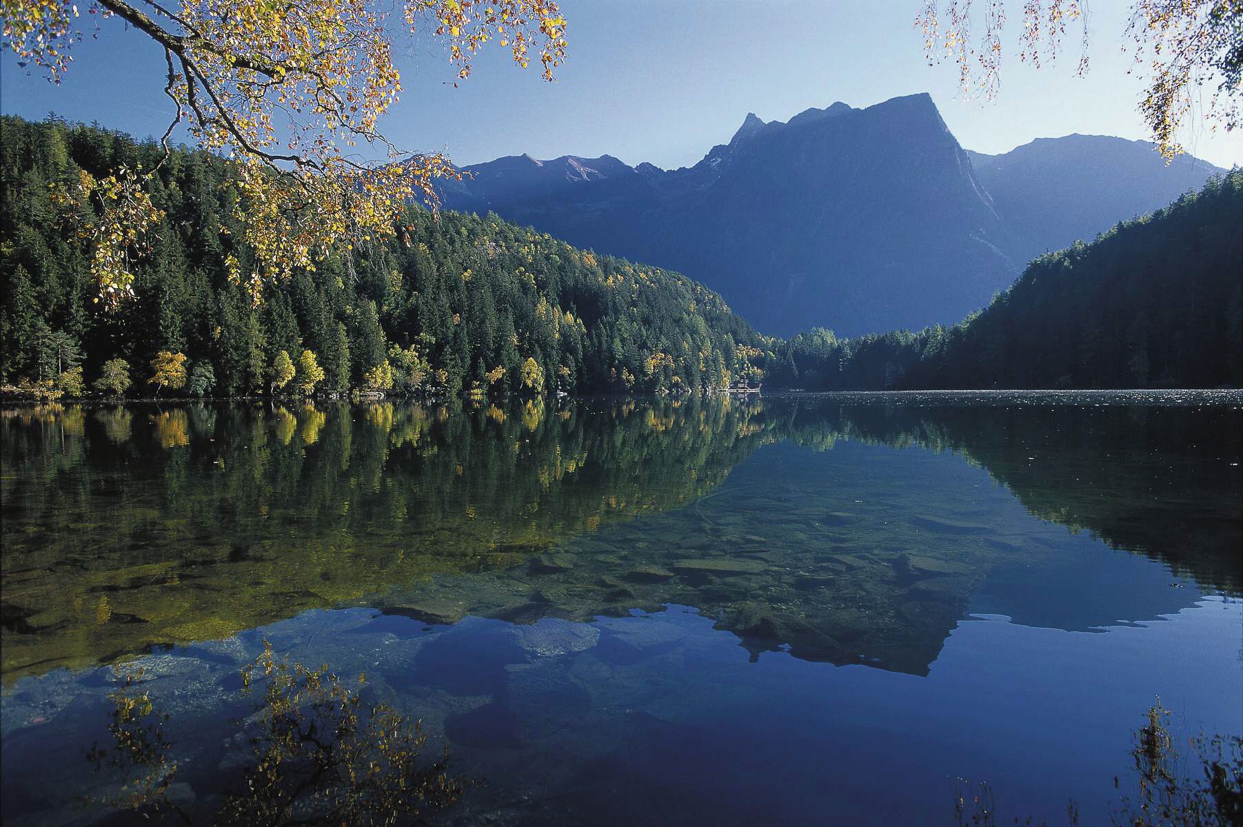 Ein ruhiger See mit klaren Spiegelungen der umliegenden Bäume und Berge.