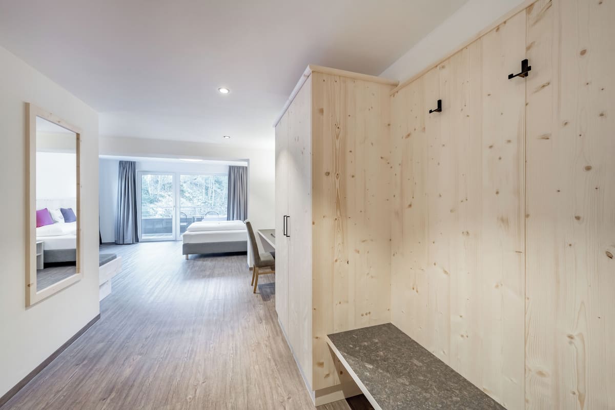 A modern hotel room with a wooden wardrobe, a bed with a view, a bench, and a large mirror.