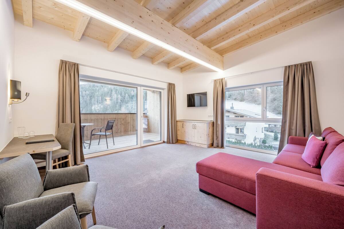 A spacious living room with a red sofa, grey armchairs, a wooden ceiling, large windows, and a view of the outdoors.