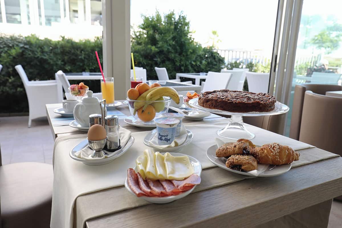 Una mesa de desayuno con platos de huevos, tocino, bollería y un pastel, además de un frutero y un vaso de zumo de naranja.