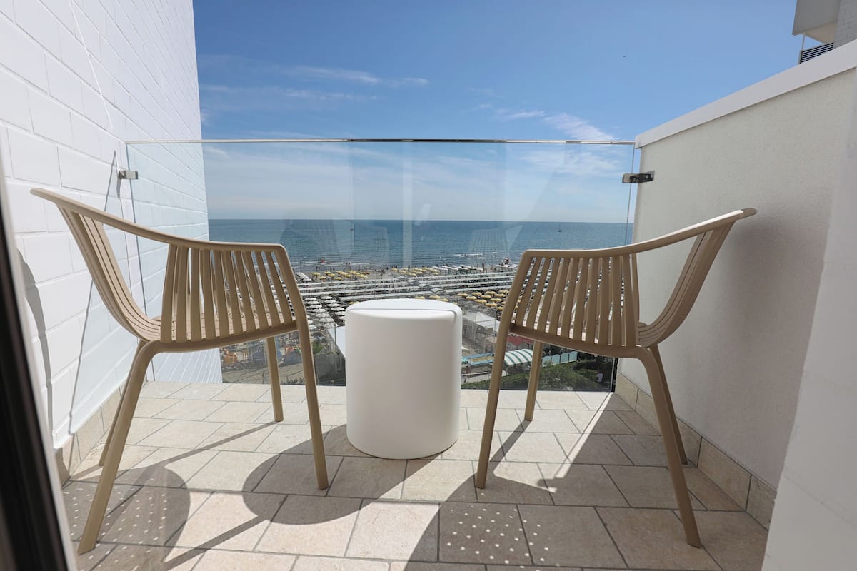 Un balcón con dos sillones de mimbre y un reposapiés blanco, con vista a una playa con sombrillas y gente.