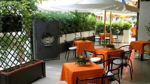 Un espacio de comedor al aire libre con mesas cubiertas con manteles naranja, sillas de metal negro y plantas en macetas.