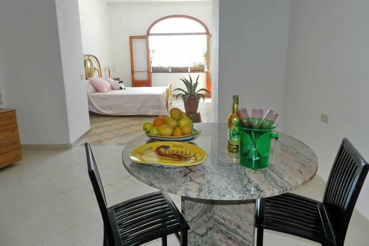 Une pièce comprenant un lit, une table à manger avec des chaises, un panier de fruits et un vase vert contenant des bouteilles.