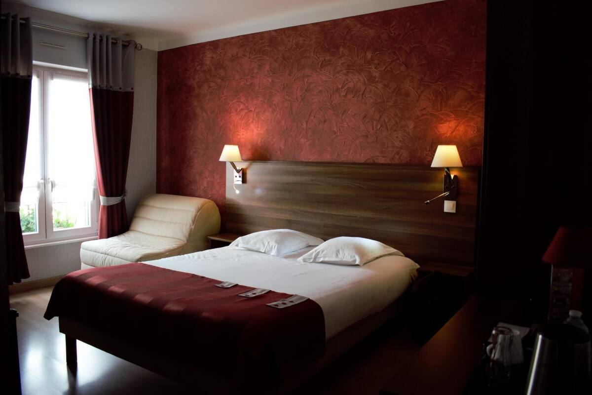 A hotel room with a bed, a chair, two wall-mounted lamps, and a window with curtains.