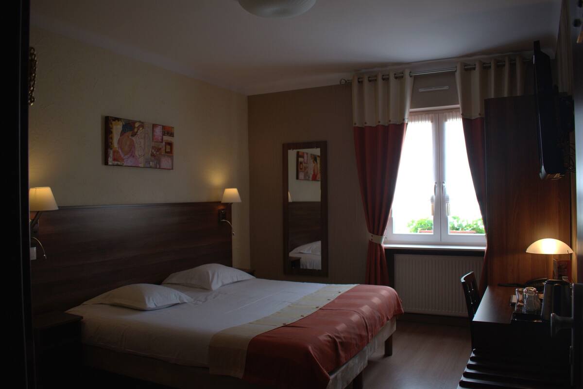 A hotel room with a large bed, two bedside lamps, a window with curtains, a wooden headboard, a mirror, and a small table with a lamp.