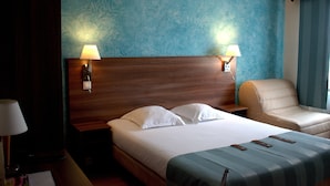A hotel room with a wooden headboard, two bedside lamps, a bed with white linens, a leather armchair, and a blue textured wall. - La Table Alsacienne (Farebersviller)