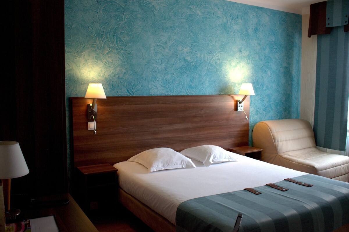 A hotel room with a wooden headboard, two bedside lamps, a bed with white linens, a leather armchair, and a blue textured wall.