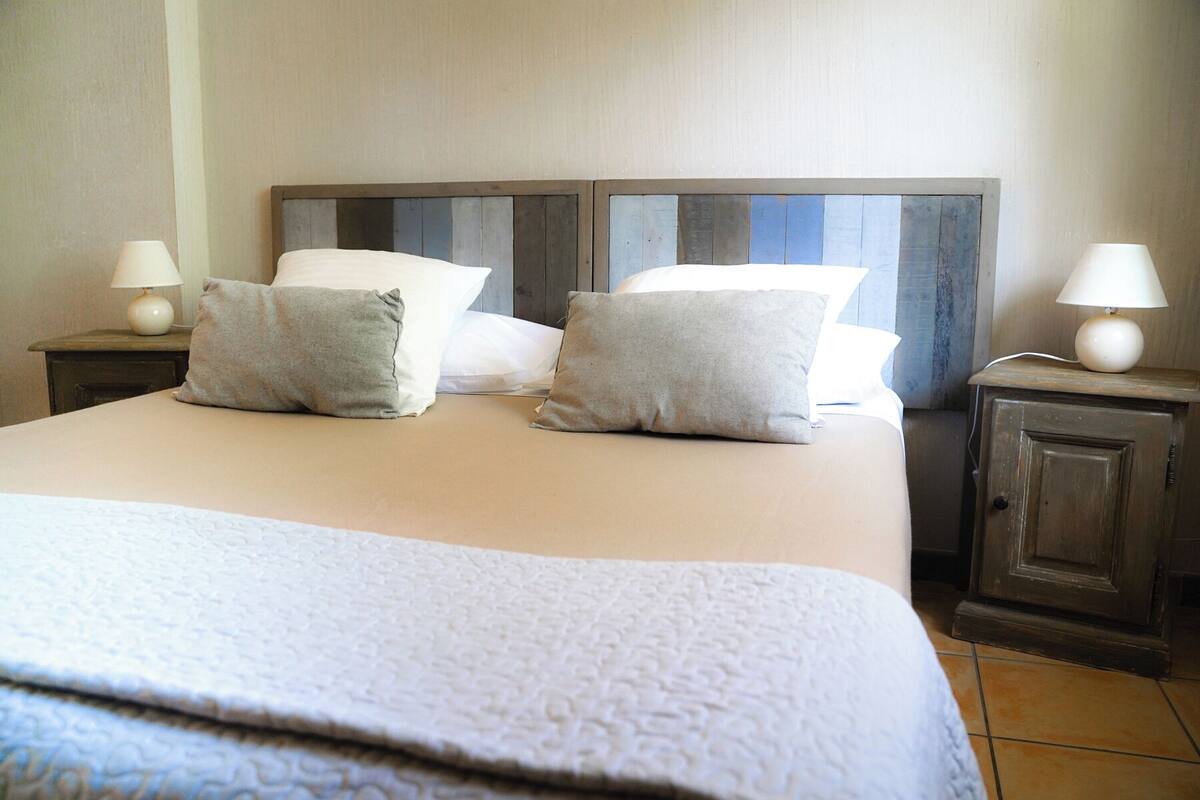A neatly made bed with a white duvet and two pillows, a wooden headboard, two bedside tables with lamps, and a tiled floor.