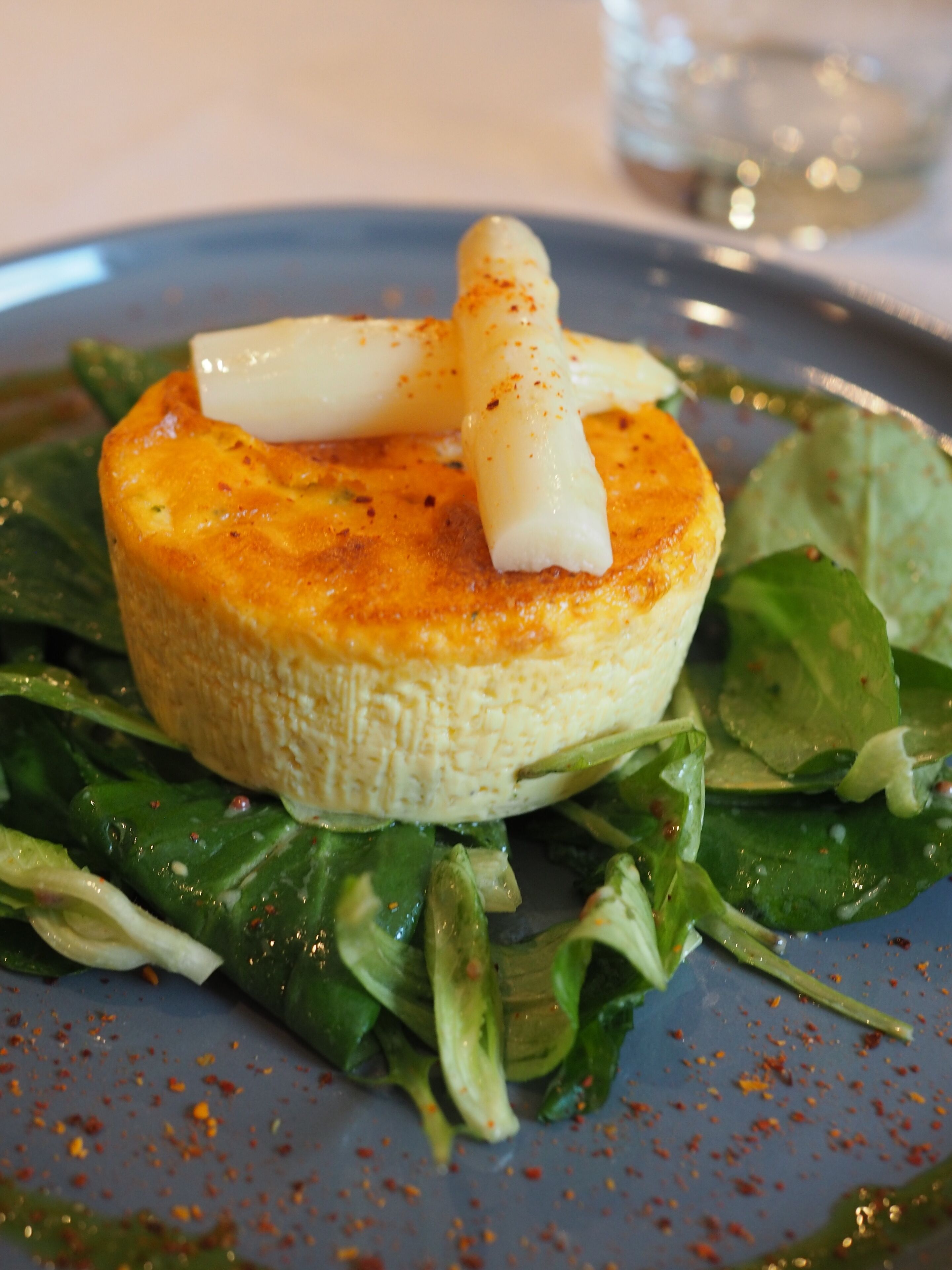 Une assiette contenant un aliment rond et de couleur brun doré, qui pourrait être une crêpe ou une omelette, surmonté d’un objet blanc et allongé, le tout saupoudré d’une pincée d’épices rouges.