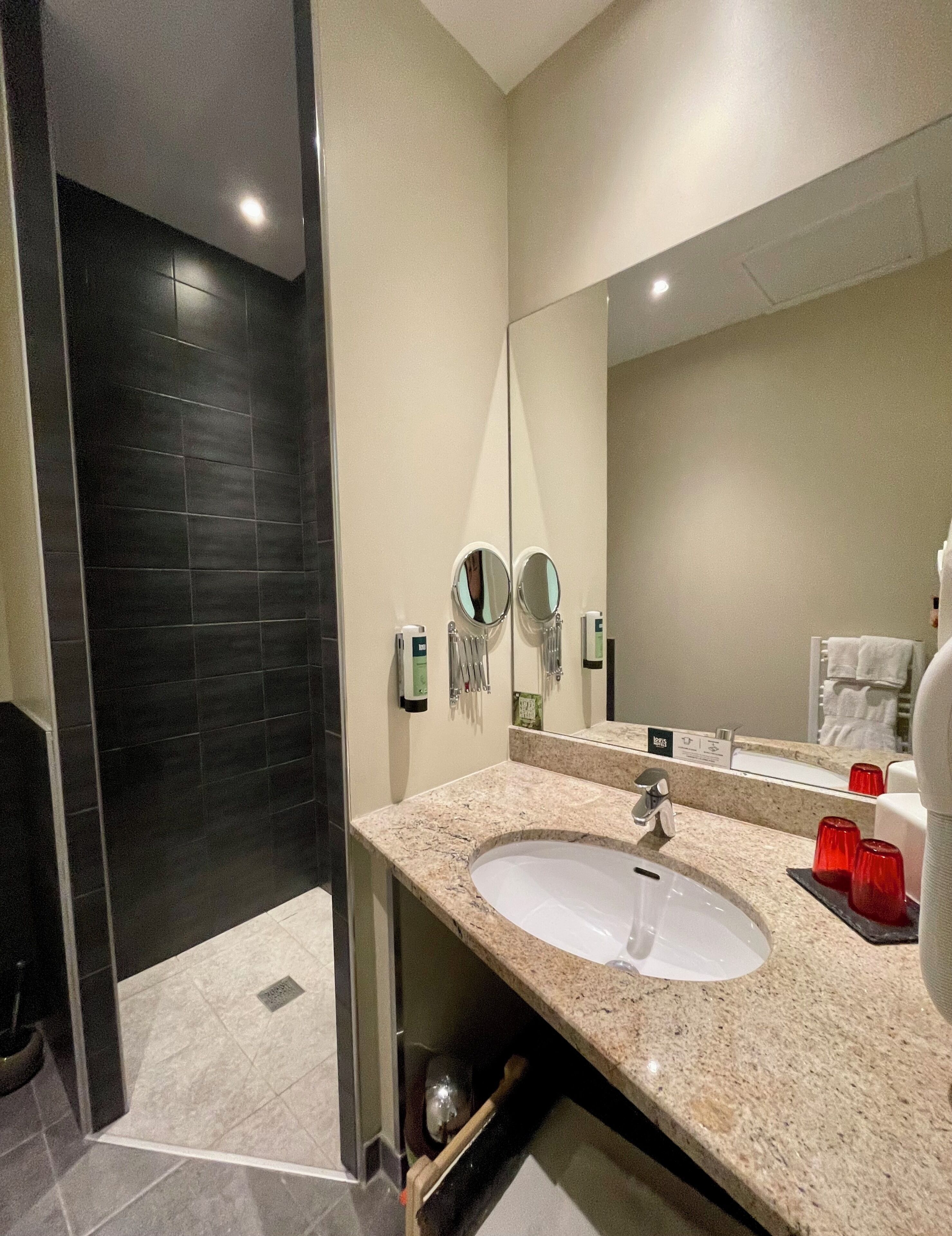 Un baño moderno con un lavamanos grande, espejos dobles y una zona de ducha con azulejos oscuros.