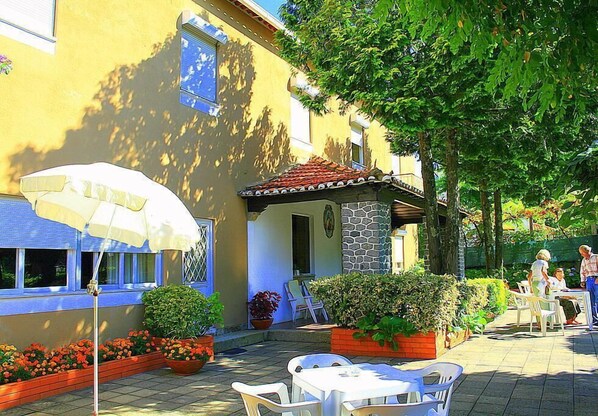 An outdoor patio with white tables and chairs, a yellow building with a stone facade, and a large tree providing shade.