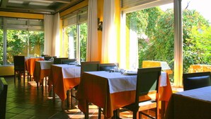 A restaurant with tables covered in white and orange tablecloths, large windows with curtains, and a view of greenery outside.