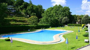 A swimming pool surrounded by grass and trees, with a few umbrellas and a flag.