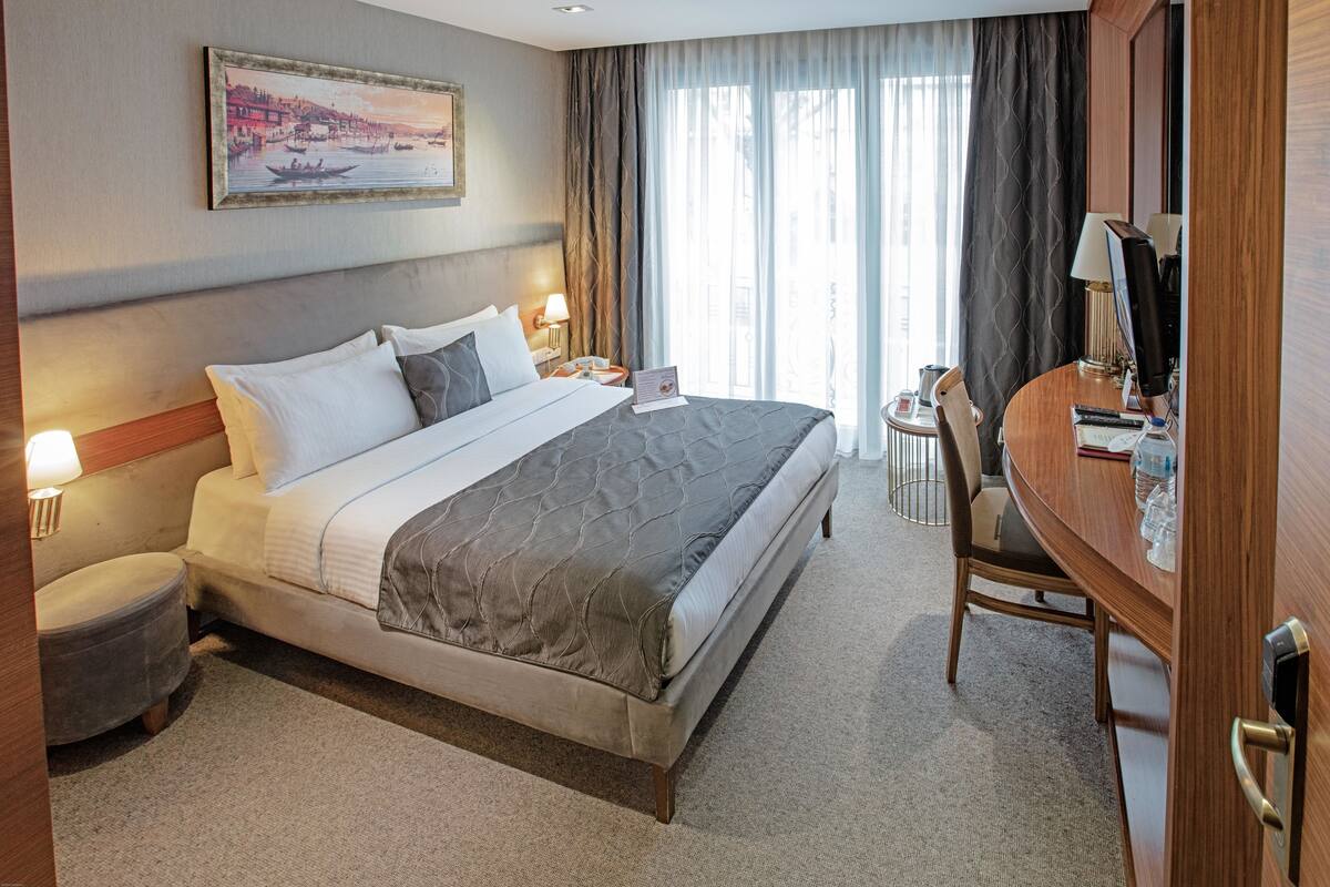 Ein Hotelzimmer mit einem großen Bett, Nachttischlampen, einem Schreibtisch mit Computer und einem Fenster mit Vorhängen.