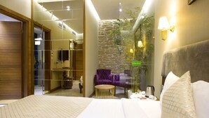 Una habitación de hotel con una cama grande, una mesita redonda de madera, un sofá púrpura, una pared de acento de piedra y una planta en maceta.