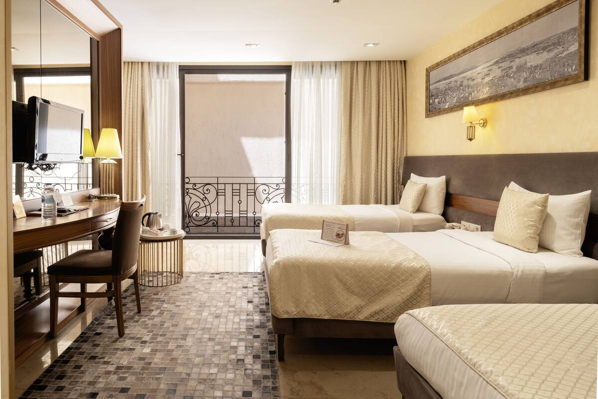 Ein Hotelzimmer mit zwei Betten, einem Schreibtisch mit Stuhl, einem Fernseher und einem Balkon mit Vorhängen.