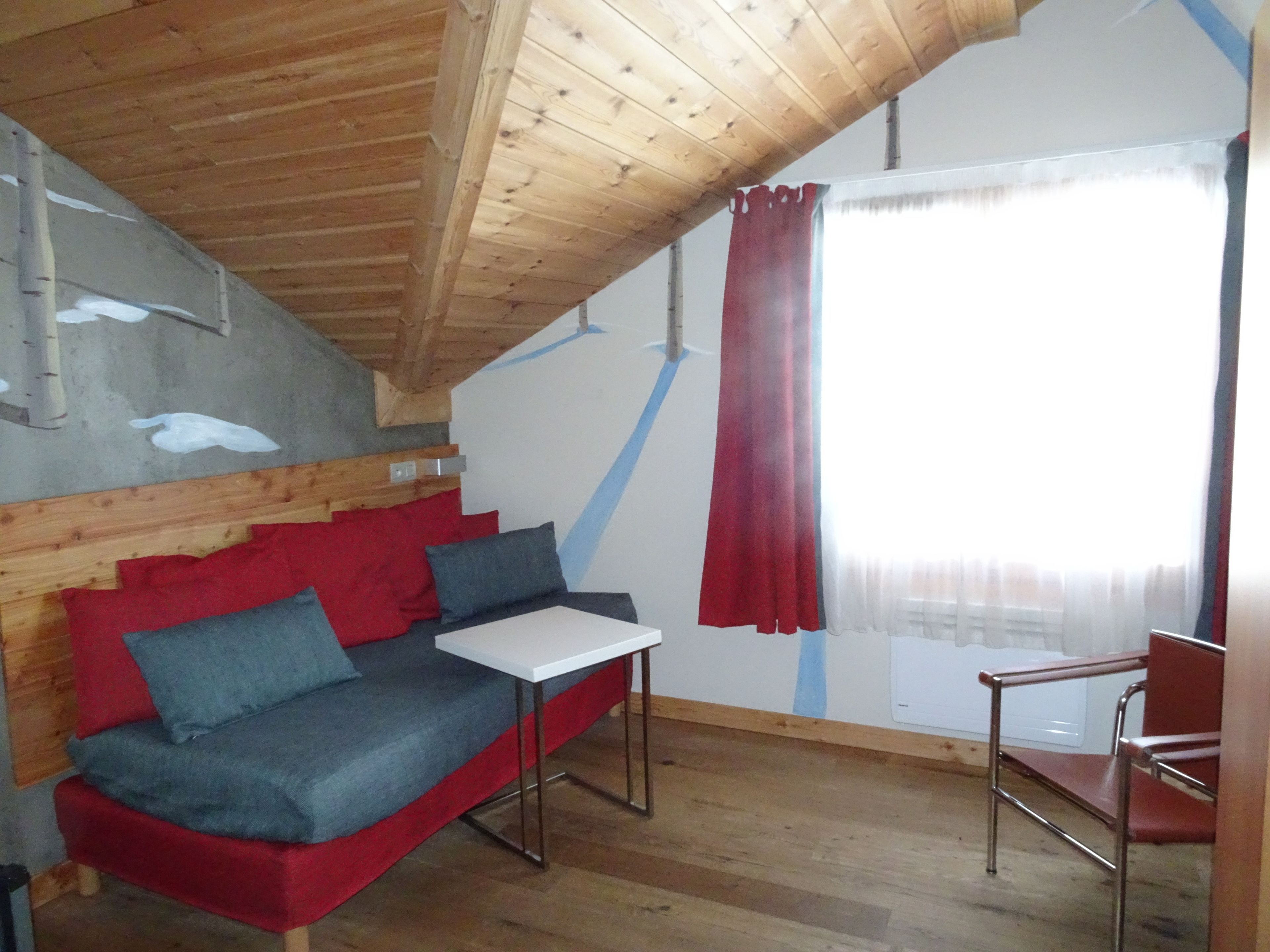 Une petite pièce accueillante avec un plafond en bois, un canapé rouge et gris, une petite table blanche et une chaise rouge.