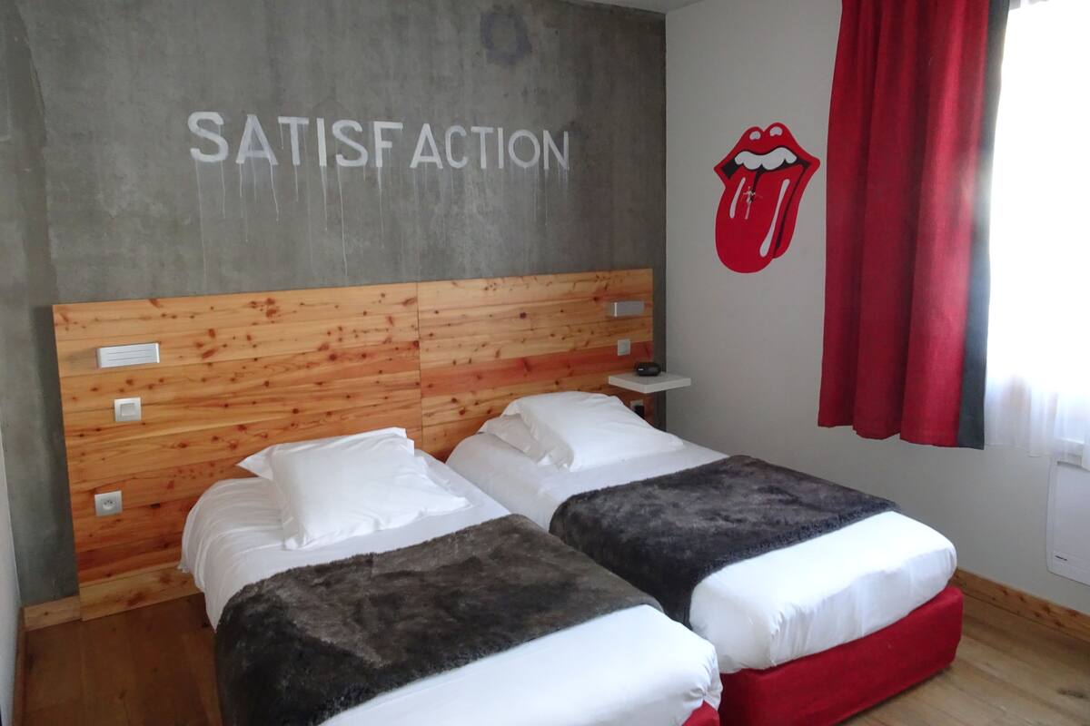 A bedroom with two beds, a wooden headboard, a wall with 'SATISF ACTION' text, a red lips mural, and a red curtain.