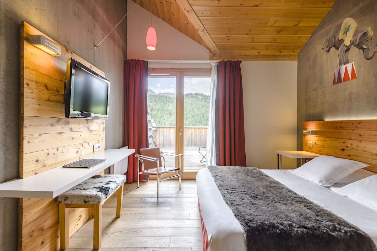 A hotel room with a wooden interior, a bed, a desk with a chair, a television, and a view of the outdoors.