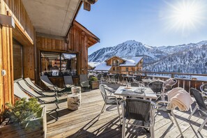 Una terraza de madera con mobiliario exterior, zona de bar y vistas a montañas nevadas.