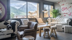 Sala de estar moderna com uma grande janela que oferece vista para as montanhas, um sofá branco, uma poltrona de couro marrom e uma mesa de centro de madeira.