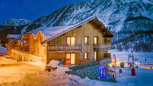 Uma cabana de madeira com varanda, um teleférico ao fundo e montanhas cobertas de neve.
