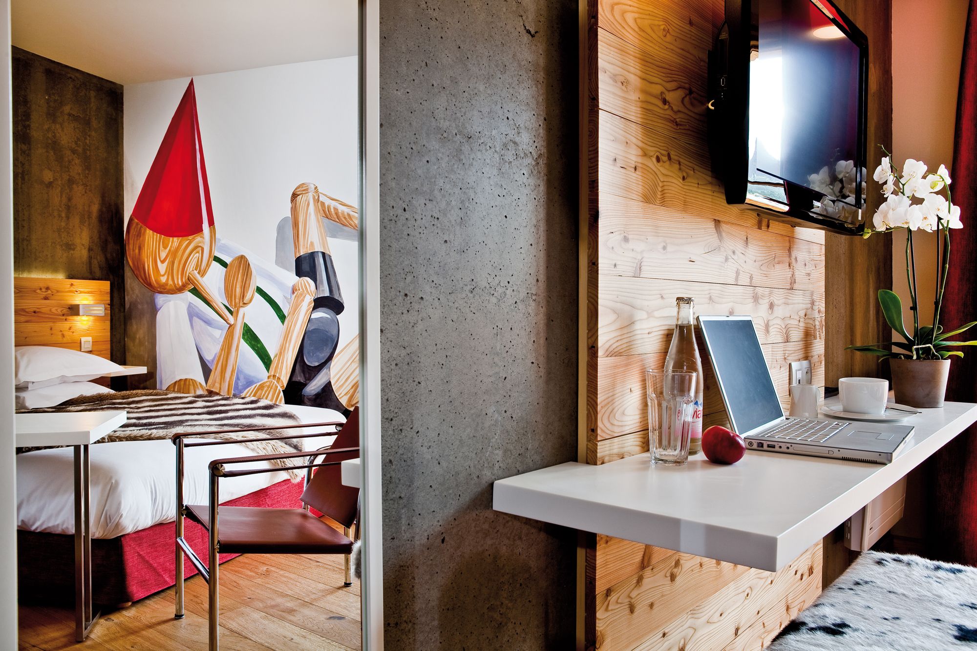 A modern hotel room with a bed, a desk with a laptop, a TV, and a potted plant.