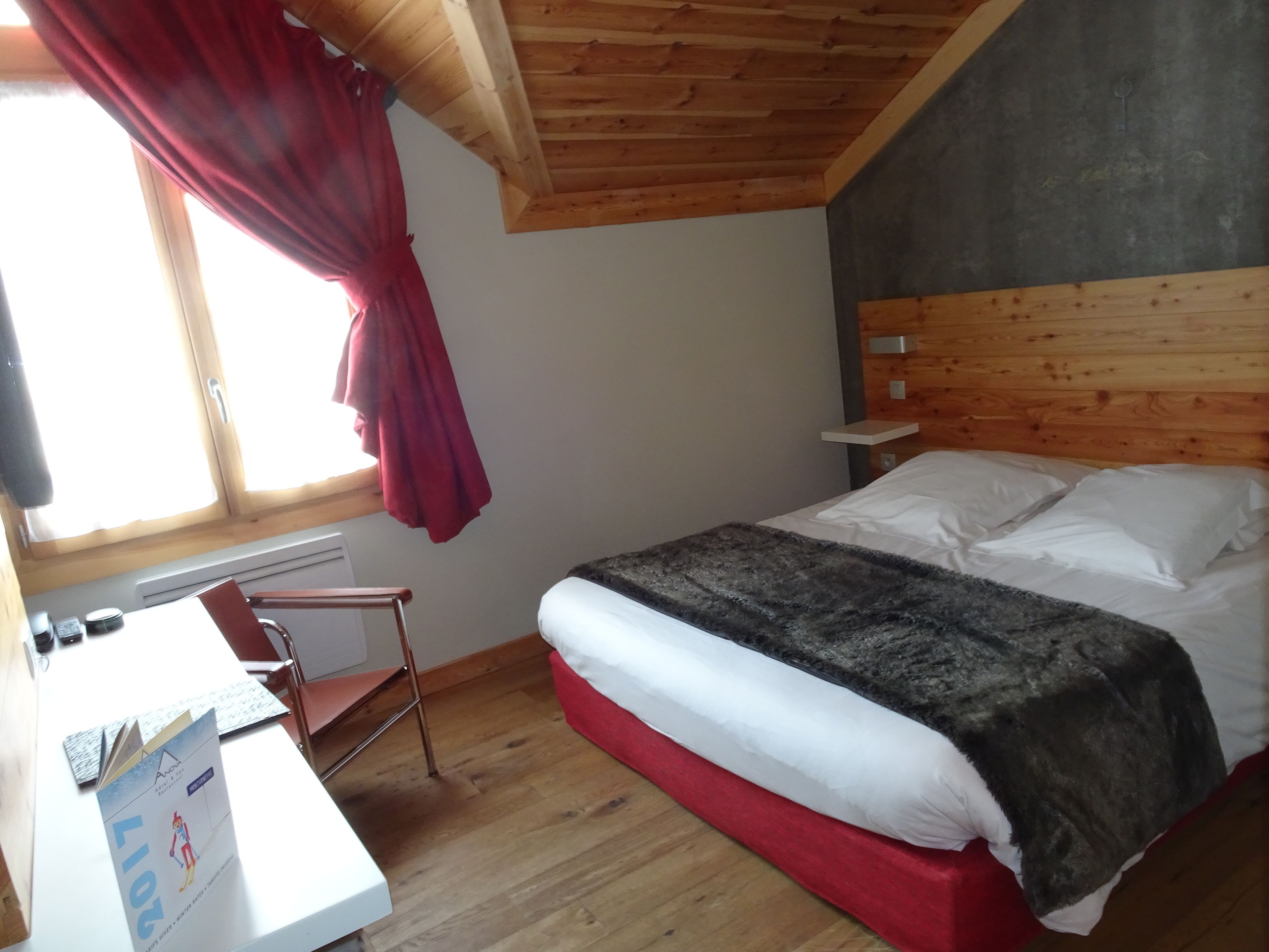 A bedroom with a wooden ceiling, a bed with white and grey bedding, a desk with a chair, and a window with red curtains.