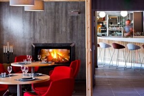 A cozy restaurant with a fireplace, red chairs, and a bar area visible through an open doorway. - Anova Hotel & Spa (Montgenevre)