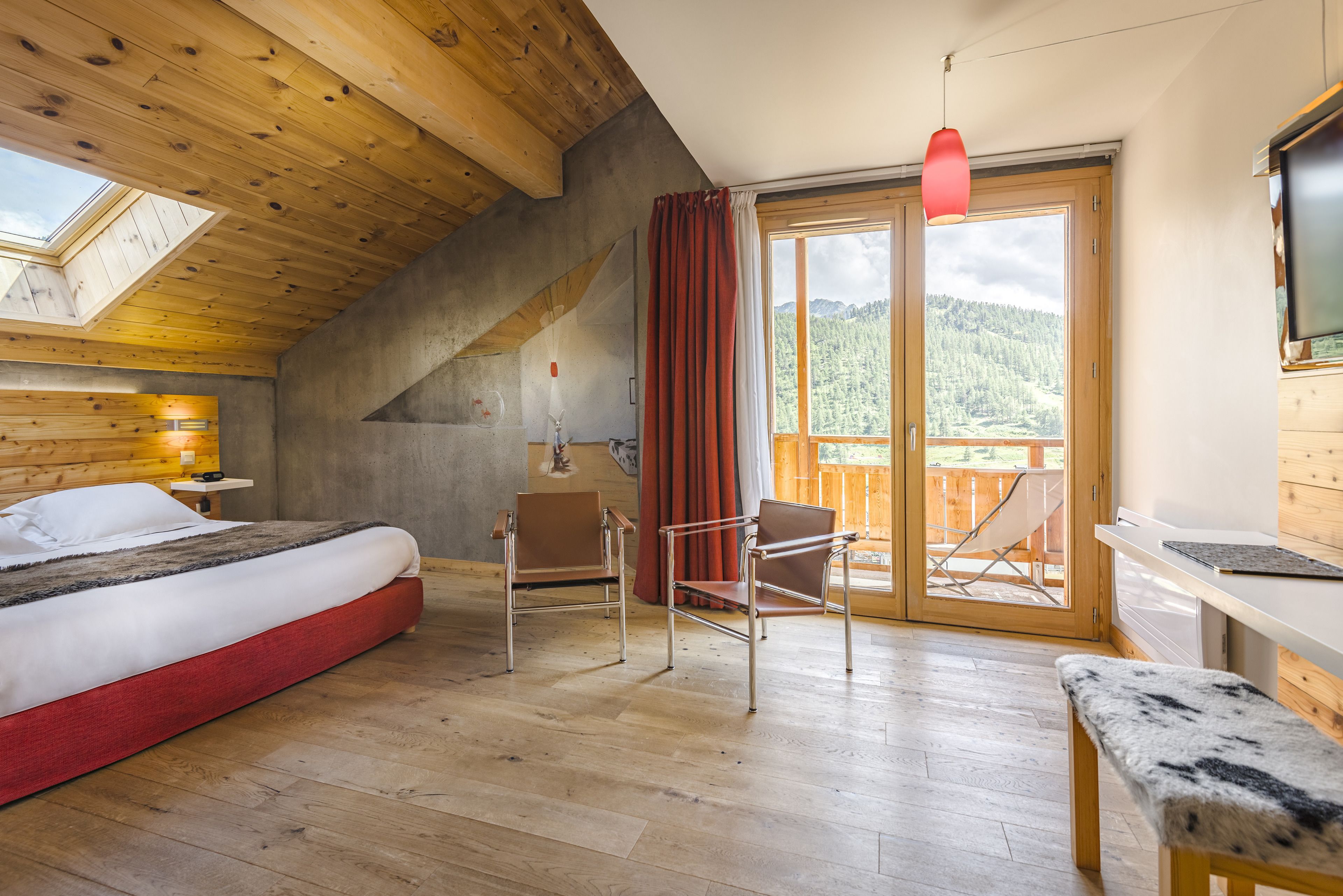 A bedroom with a bed, a desk, a chair, a bench, a red hanging light, and a sliding glass door leading to a balcony with a view of a forested mountain.