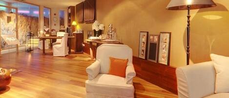 A spacious room with a wooden floor, a white armchair with an orange pillow, a white sofa, and a lamp.