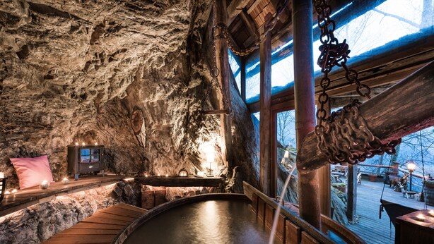 Un interior rústico con pared de piedra, asientos de madera, un jacuzzi y una terraza de madera con mobiliario exterior visible a través de una ventana.
