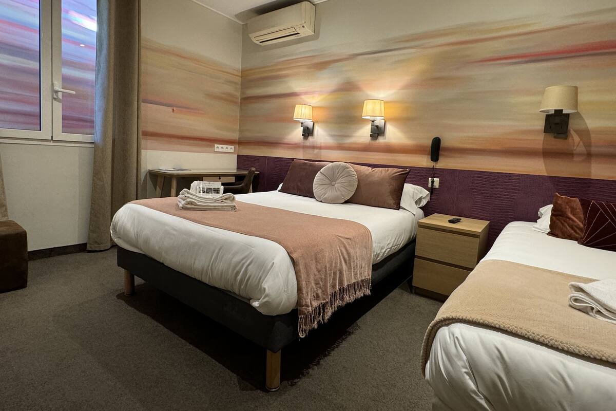 A hotel room with a large bed, a smaller bed, a desk, and a chair. There is an air conditioning unit on the wall and two wall-mounted lamps.