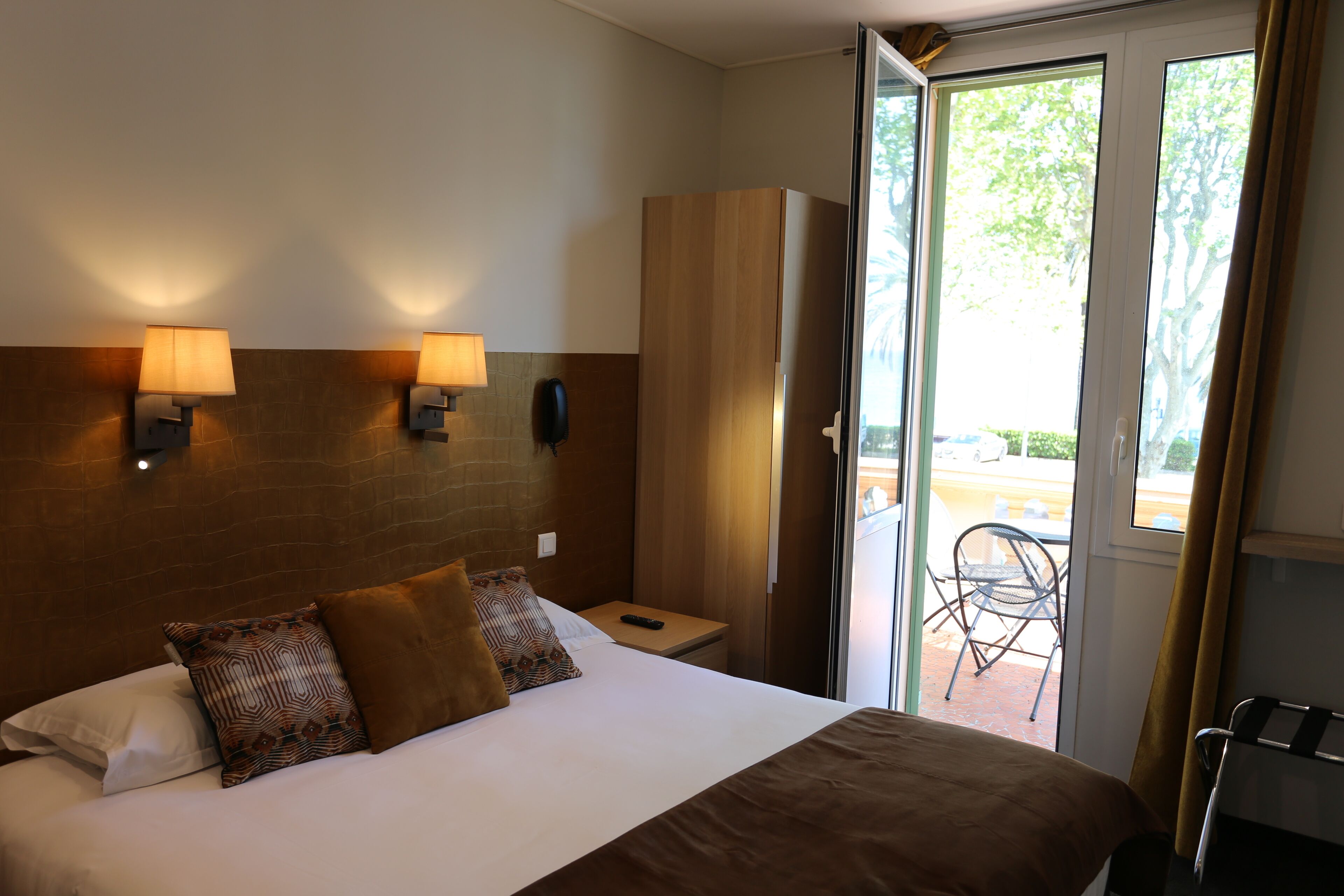 Une chambre d’hôtel comprenant un lit, deux lampes de chevet, une armoire, une petite table et une chaise, le tout à proximité d’une porte-fenêtre coulissante donnant sur un espace extérieur.