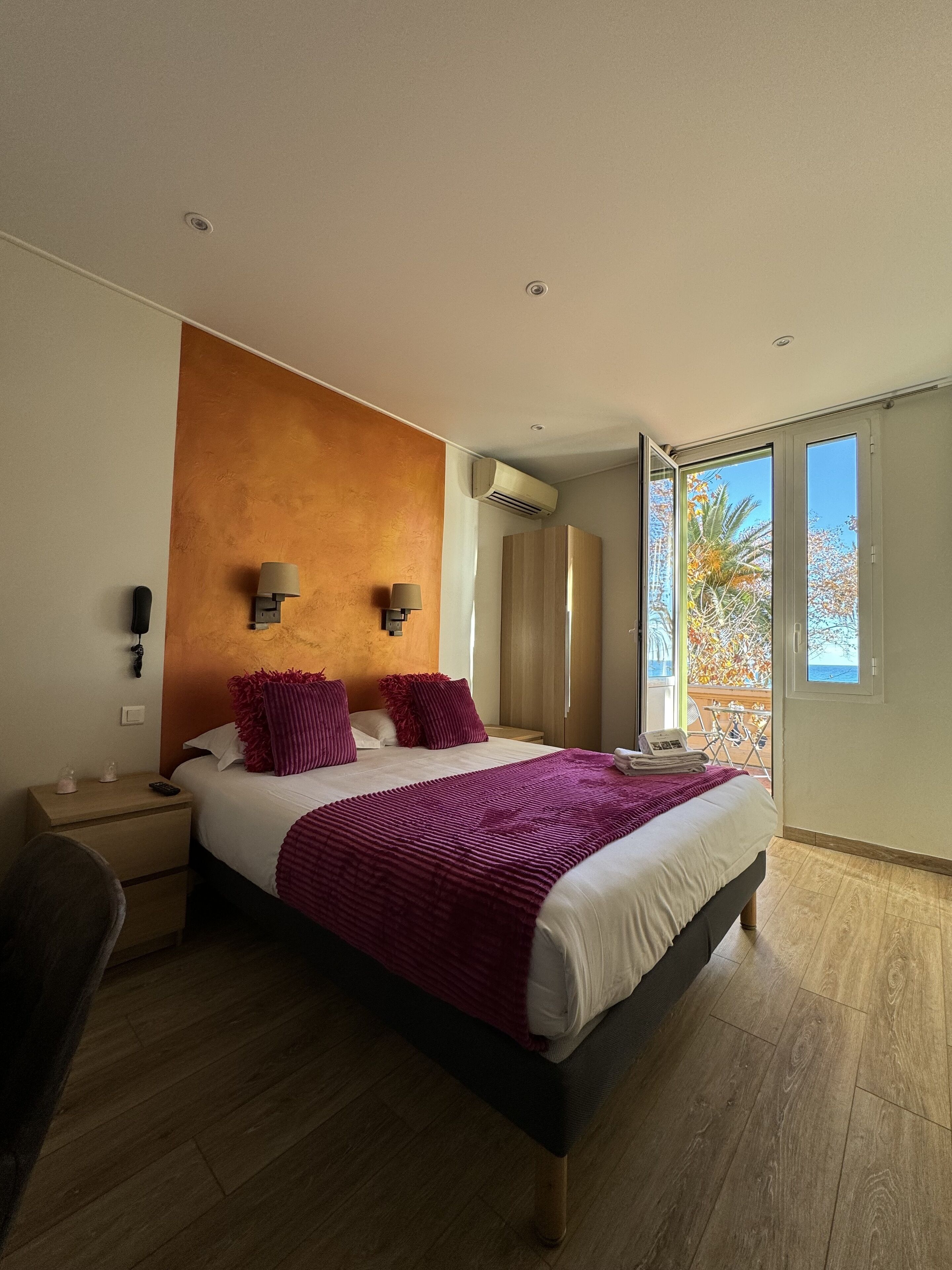 A modern hotel room with a large bed, two bedside lamps, a nightstand, a desk, and a window with a view of trees.