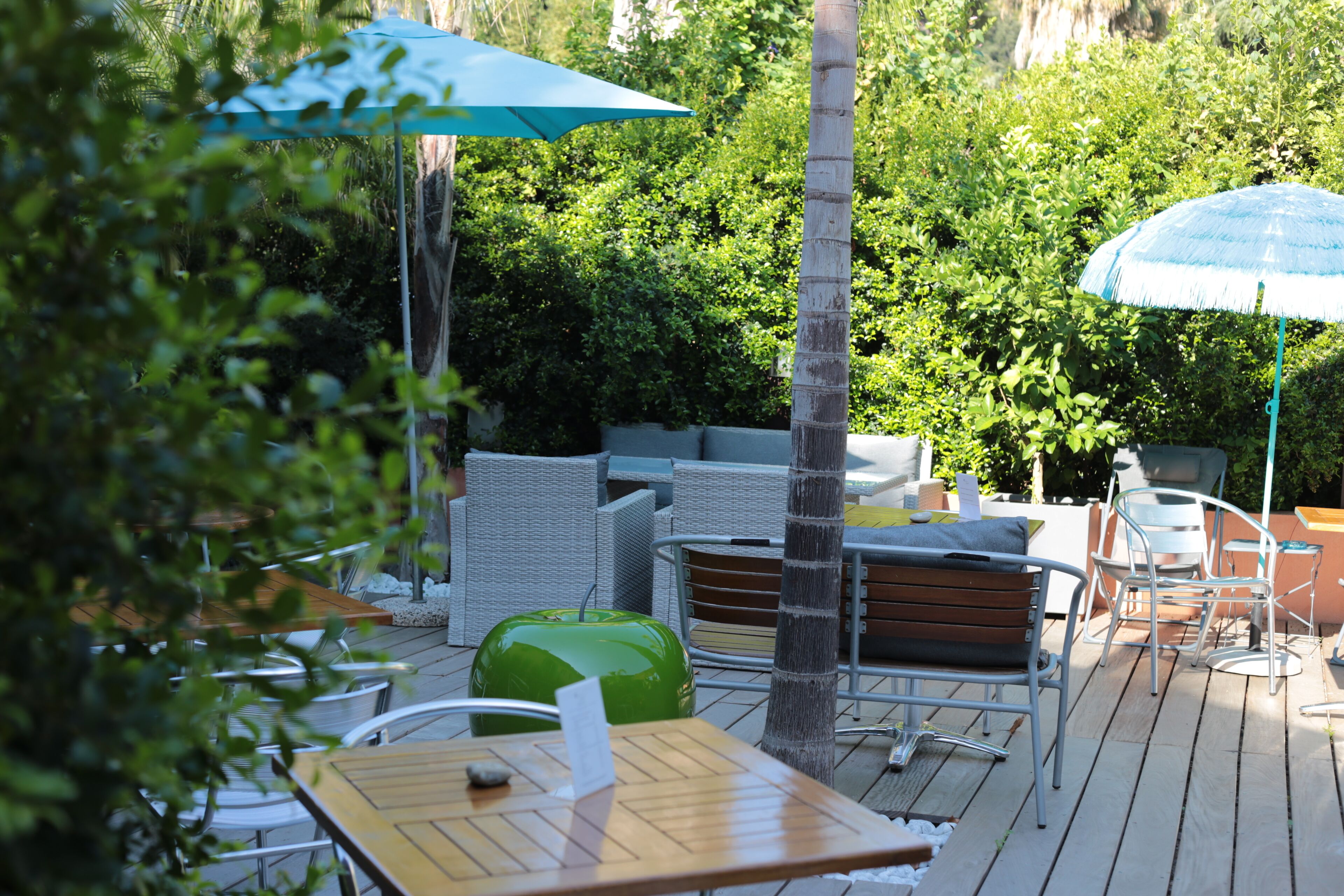 An outdoor patio area with wooden tables and chairs, a green suitcase, and two umbrellas providing shade.