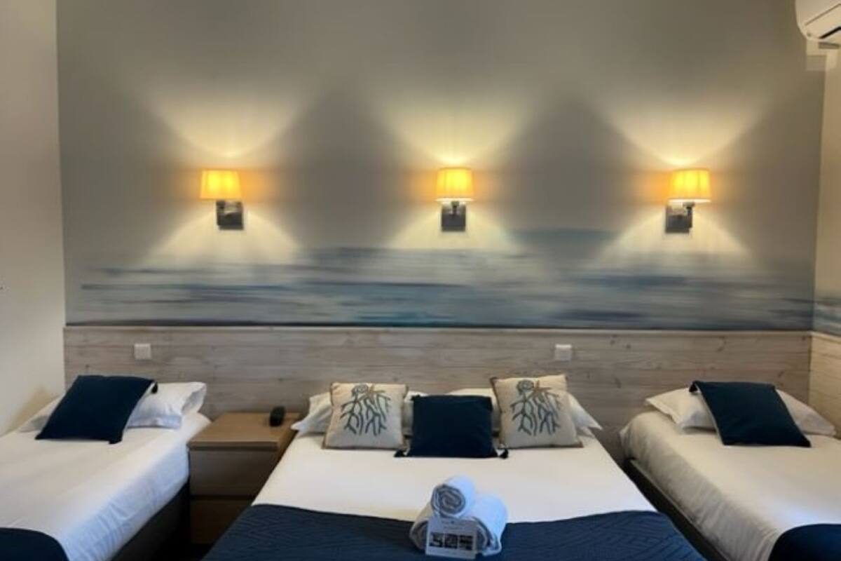 A hotel room with three beds, each with a pillow and a blanket. There are wall-mounted lamps on the ceiling, and a painting of a seascape on the wall behind the beds.
