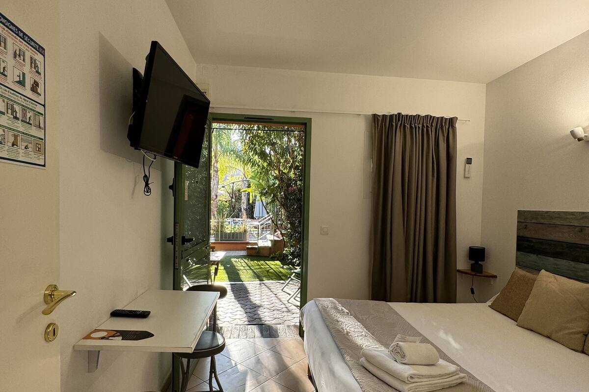 A hotel room with a bed, a desk with a chair, a television mounted on the wall, and a view of the garden through the door.