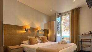 Habitación de hotel con una cama grande, dos lámparas de pared, vista al mar a través de una puerta corredera, una mesita con una silla y una estantería con objetos.