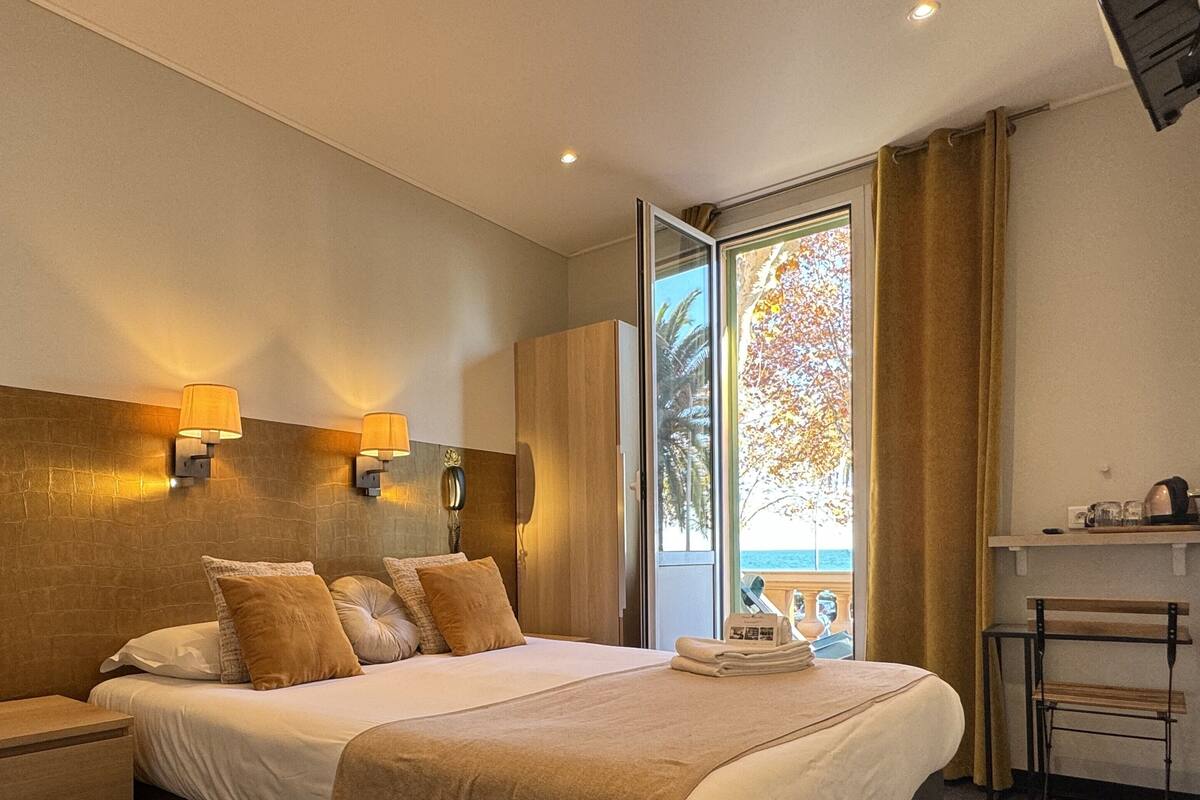 A hotel room with a large bed, two wall-mounted lamps, a view of the sea through a sliding door, a small table with a chair, and a shelf with items.