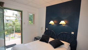 Habitación de hotel con cama, dos lámparas de pared, ventana con vista a palmeras y una mesita de noche.