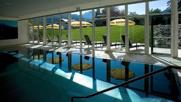 Una piscina con pareti di vetro trasparente, lettini prendisole e vista su una zona verde con ombrelloni.