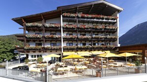 Un edificio a più piani con balconi decorati con fiori, sedute all'aperto sotto ombrelloni gialli, e montagne sullo sfondo.