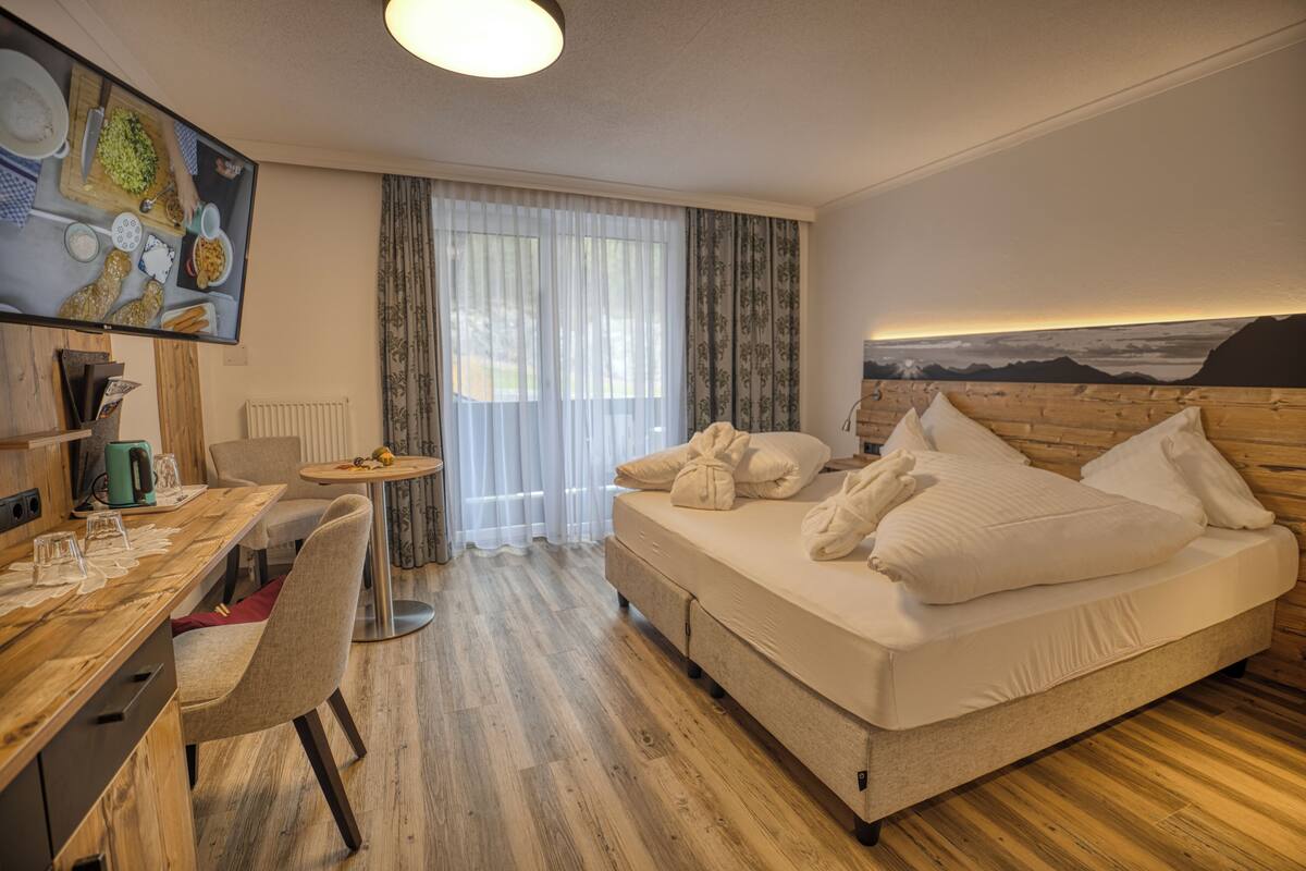 A hotel room with a wooden floor, a bed with white linens, a desk with a chair, a small table with a vase, and a painting on the wall.