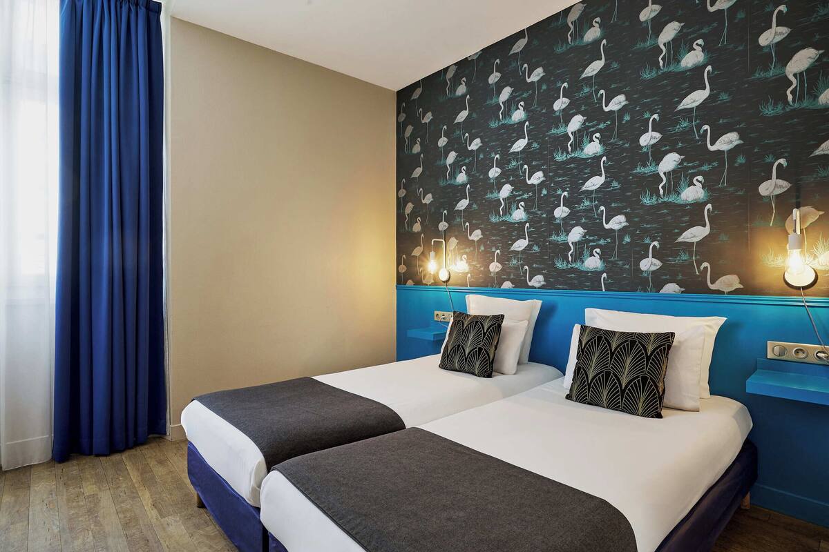 Ein Hotelzimmer mit zwei Betten, einem blauen Kopfteil, einer Wand mit Vogelmotiv und einem Fenster mit blauen Vorhängen.