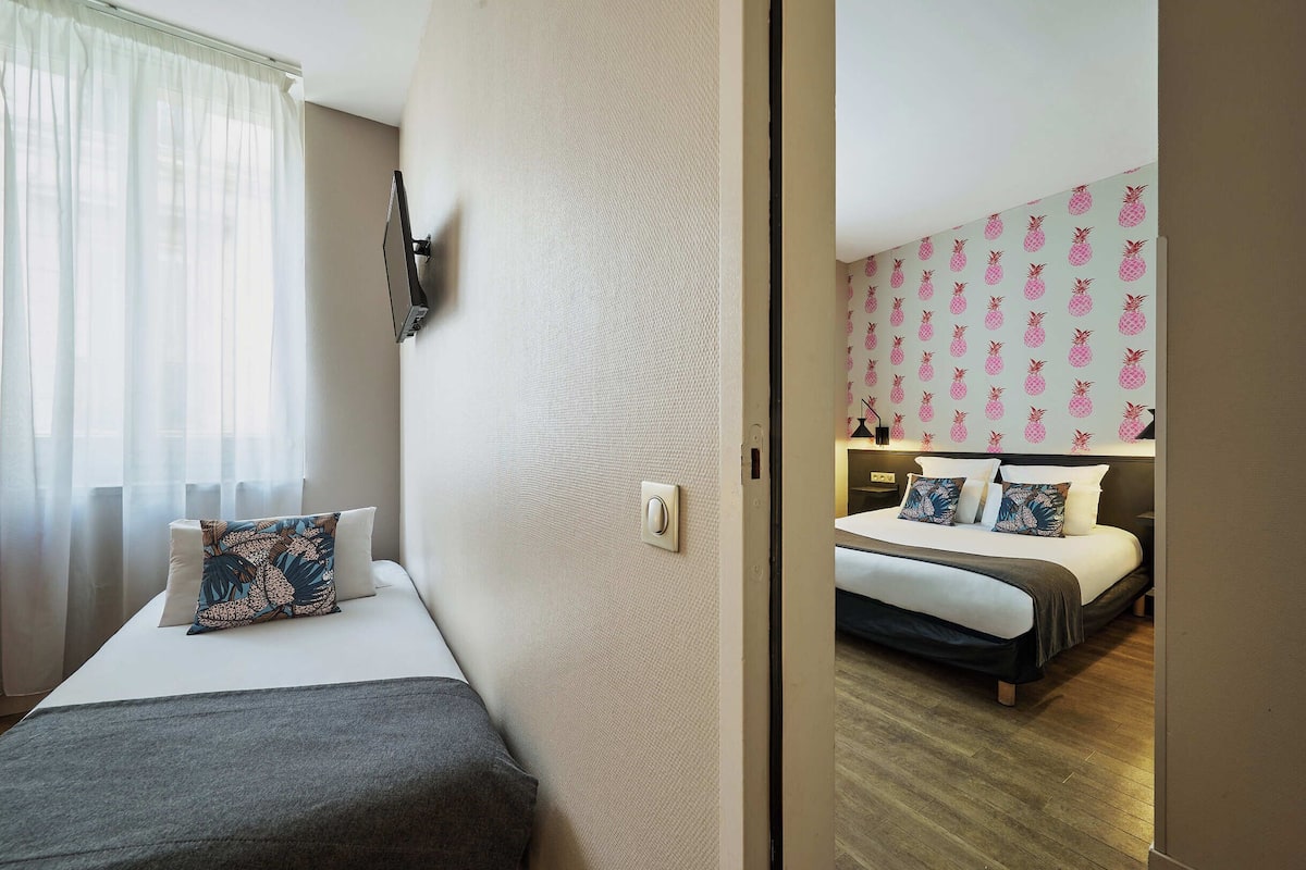 Ein Hotelzimmer mit Bett, Nachttisch, Flachbildfernseher und Fenster mit leichtem Vorhang.