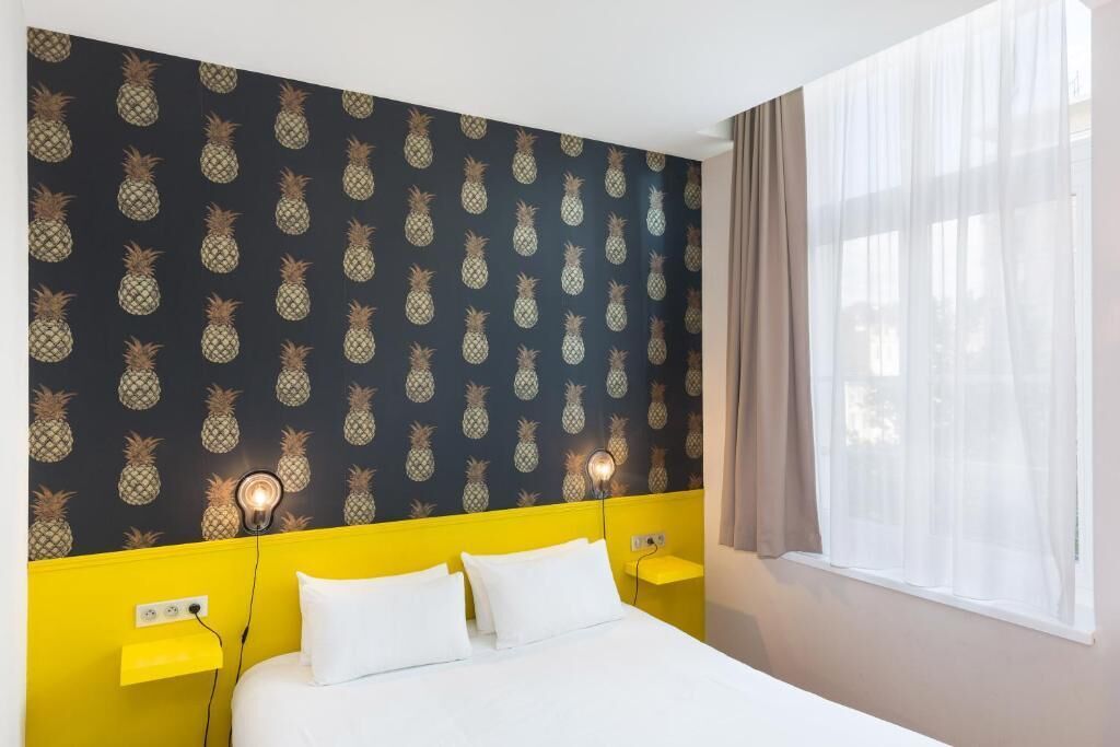 Une chambre d’hôtel avec un lit, une tête de lit jaune et un papier peint à motifs d’ananas.