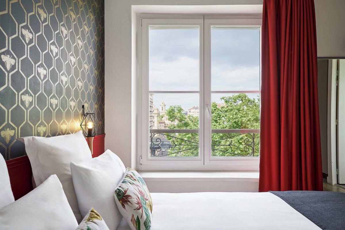 Ein Hotelzimmer mit Bett, Fenster mit rotem Vorhang und Blick auf die Stadt, sowie einer Wand mit Muster.